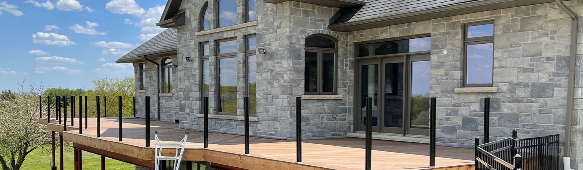 Custom wood deck surrounded by Canadian-made glass railing in backyard of large grey brick house.